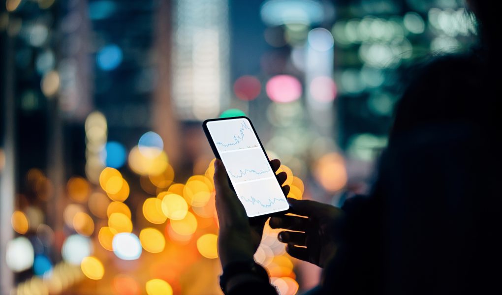 Woman checking financial stock market analysis on smartphone in city, with illuminated city street light and urban skyscrapers as background at night