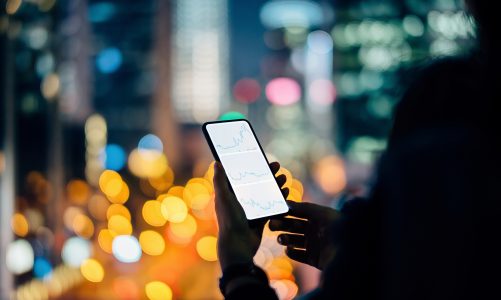 Woman checking financial stock market analysis on smartphone in city, with illuminated city street light and urban skyscrapers as background at night